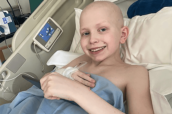 Picture of young boy in hospital bed with IV drip in his hand. Smiling widely at the camera.
