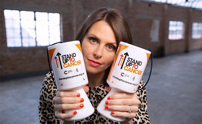 Ellie Taylor holding up two Stand Up To Cancer donation pots in front of a brick wall 