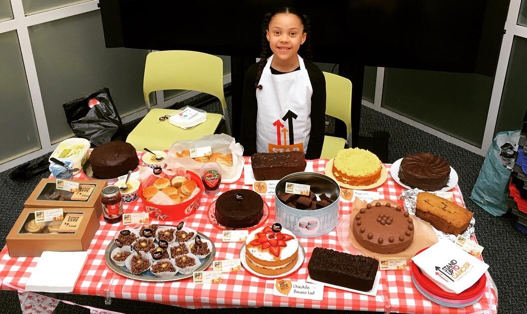 Aliza standing in front of a table of cakes for her Stand Up To Cancer baking event