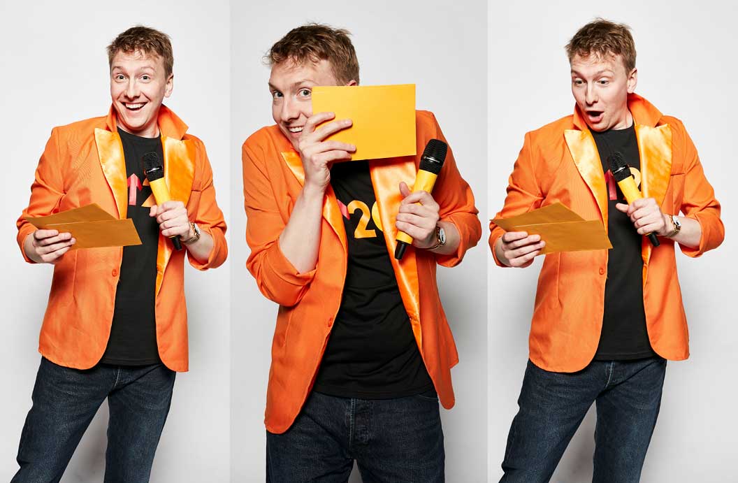 Joe Lycett holding a microphone and quiz cards in a orange jacket, smiling and laughing