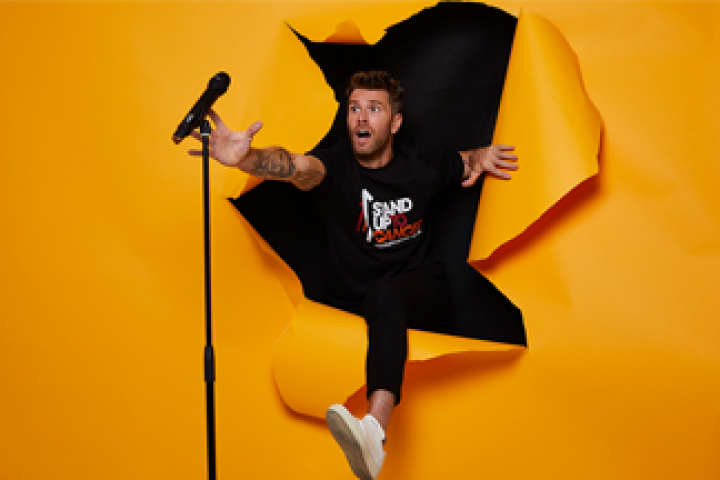 Comedian Joel Dommett breaking through an orange screen to reach his microphone