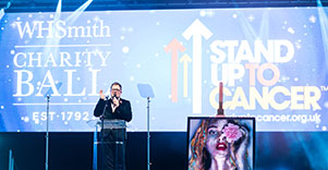 A gala host stands in front of a screen with the words, 'WH Smith Gala' and the stand up to cancer logo.