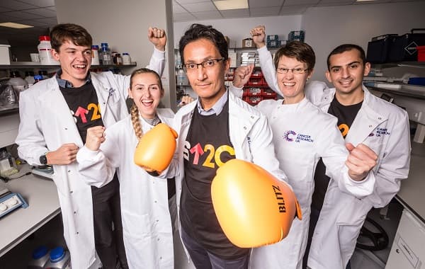 5 scientists in cancer research and stand up to cancer gear cheer for the camera with the middle man wearing orange boxing gloves.